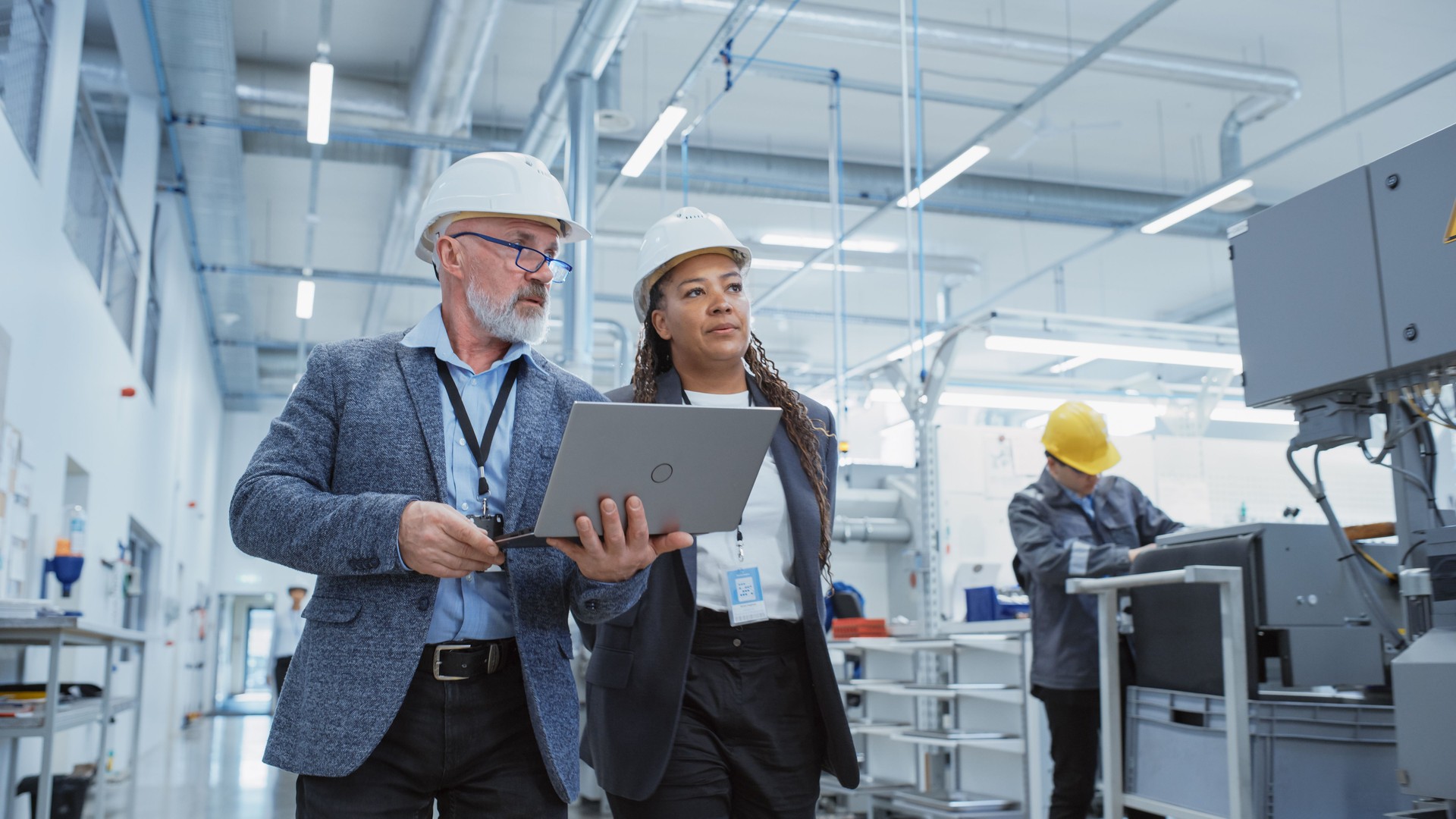Two Professional Heavy Industry Engineers Wearing Hard Hats at Factory. Walking and Discussing Industrial Machine Facility, Working on Laptop. African American Manager and Technician at Work.